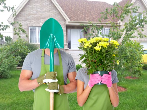 A garden scene featuring two individuals standing on a well-maintained lawn in front of a suburban house with a brick facade and large windows. The person on the left, wearing a gray t-shirt, a green apron, and yellow gardening gloves, holds a green garden shovel with a metallic blade, positioned upright. The person on the right, also in a gray t-shirt and pink gardening gloves, holds a pink pot containing a vibrant cluster of yellow flowers with green foliage, obscuring their face. The background includes a neatly trimmed hedge, a young tree with light green leaves, flowering plants, and a paved pathway leading to the house, creating a lush and orderly outdoor environment typical of Hackney gardens. The scene suggests outdoor maintenance or gardening activities, emphasizing lawn and plant care, consistent with services offered by Lawn Mowing Hackney in the local area.