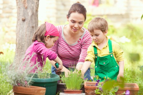 A woman with dark hair tied back and dressed in a pink and white striped top is gardening outdoors in a front or backyard with two young children, a girl wearing a pink headscarf and purple top, and a boy in a yellow shirt and green overalls. They are engaged in planting or potting activities around a collection of various containers, including terracotta and plastic pots, with flowers and green plants visible. The garden area features a lush green lawn, some shrubs, flowering plants, and a large tree trunk in the background, with soft natural lighting suggesting a sunny day. The scene captures an outdoor space maintained with care, indicative of garden and lawn maintenance services provided by Lawn Mowing Hackney, enhancing the property's landscaping and outdoor appeal with attention to plant care and garden organization.