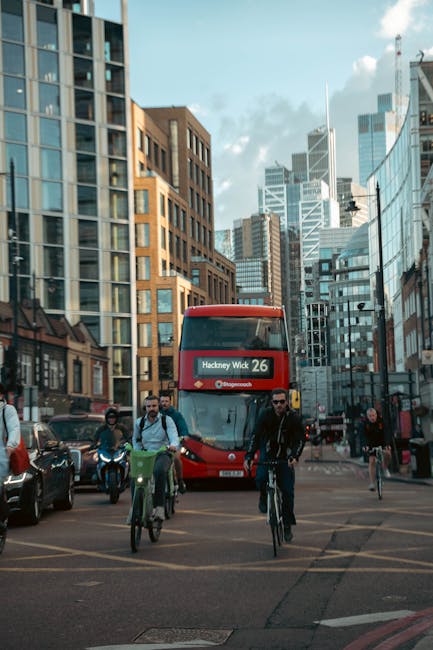 A busy urban scene in Hackney Wick featuring a red double-decker bus on the road displaying the route number 26 and destination 'Hackney Wick.' In the foreground, three cyclists navigate the street; o