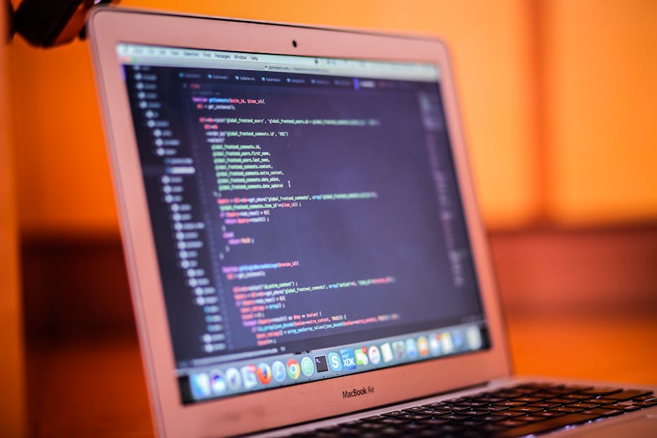 Close-up image of a pink MacBook Air with a blurred background in warm orange tones. The screen displays a code editor with colorful lines of programming code, indicating the user is working on a codi